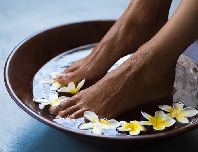 Woman soaking feet in bowl of water with floating frangipani flowers at spa.